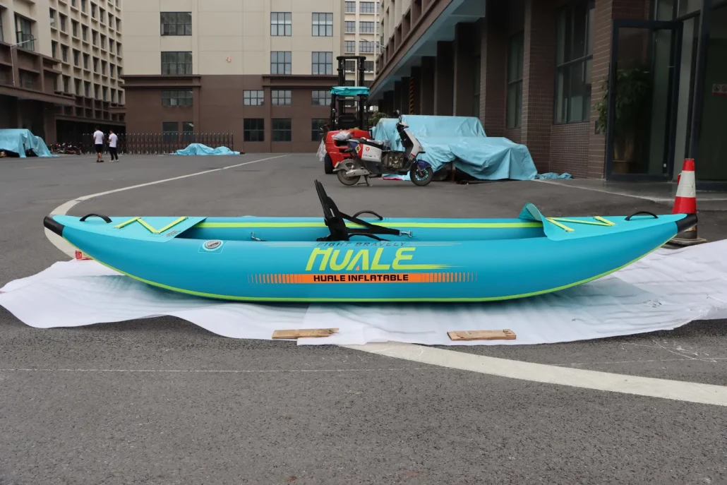 A blue inflatable kayak labeled 'Huale Inflatable' displayed on a cloth in a courtyard with a industrial building backdrop.
