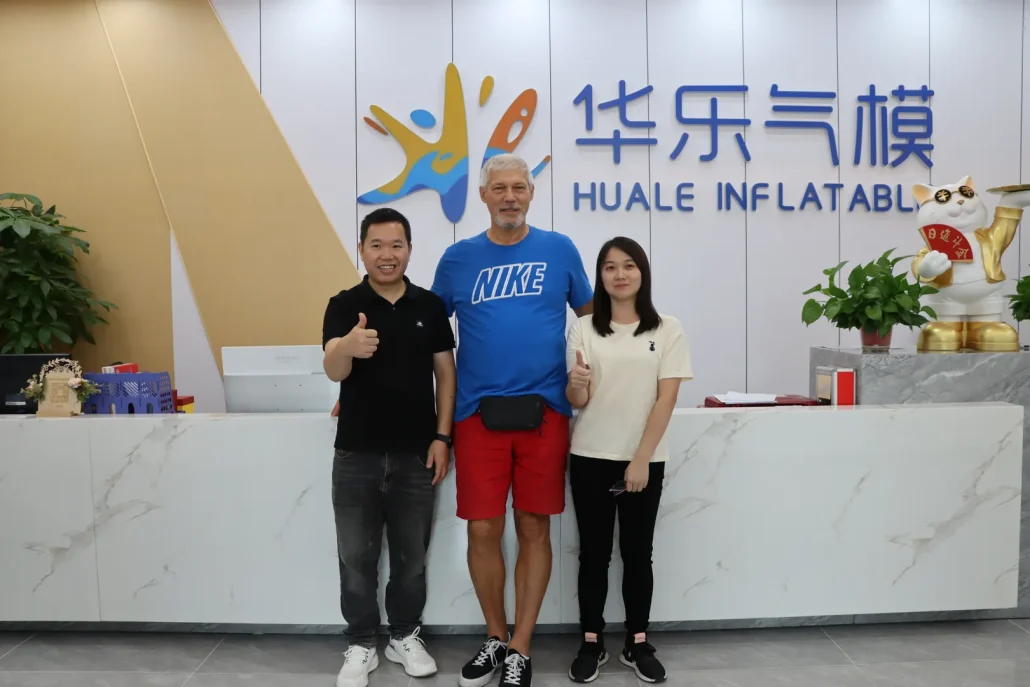 Three people stand together inside a modern office space with signage 'Huale Inflatable' on the wall and a decorative object on the desk nearby.