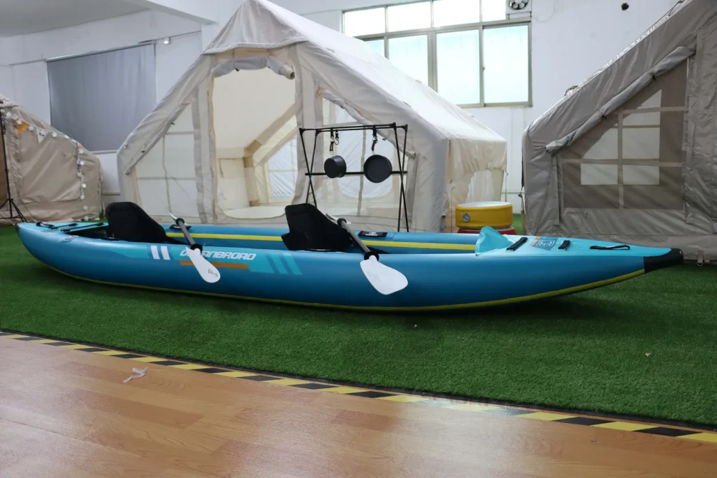 A blue and green kayak placed indoors in front of large tents with artificial grass underneath.