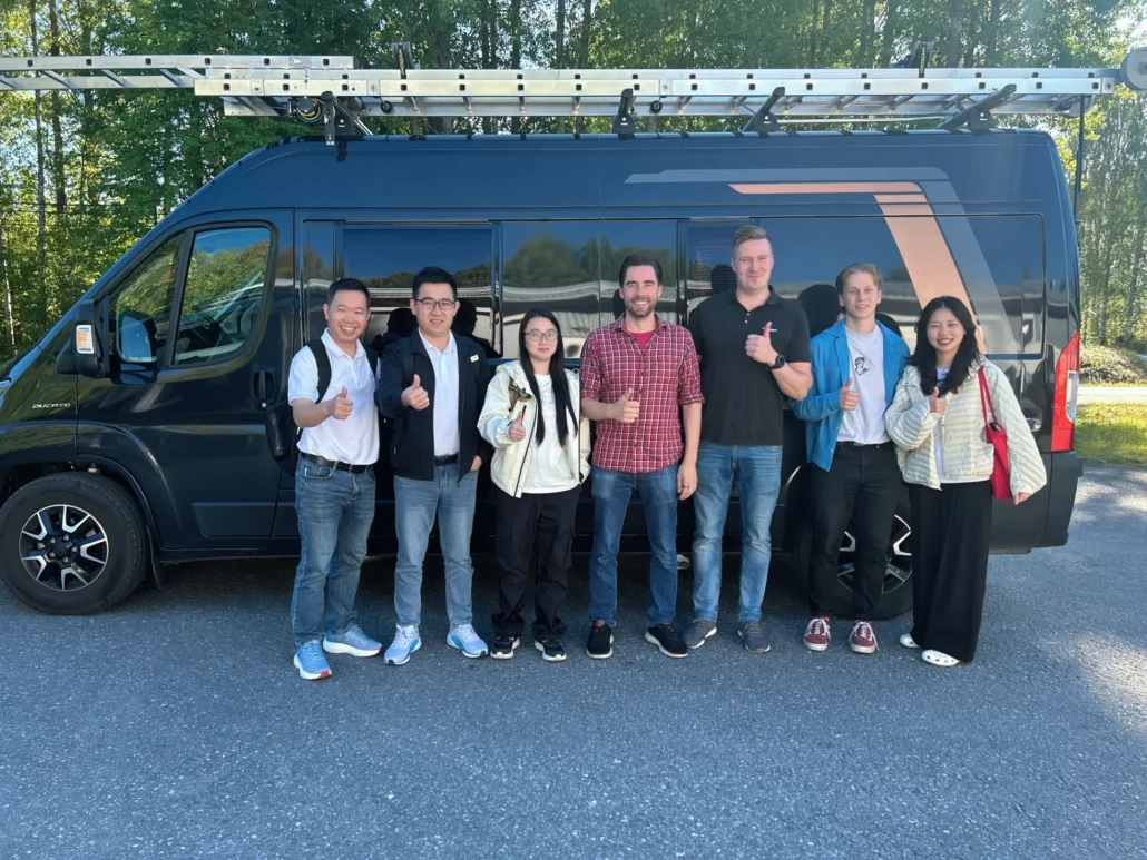 Six people standing in front of a dark blue van with a rooftop ladder, giving thumbs up gestures against a backdrop of trees.