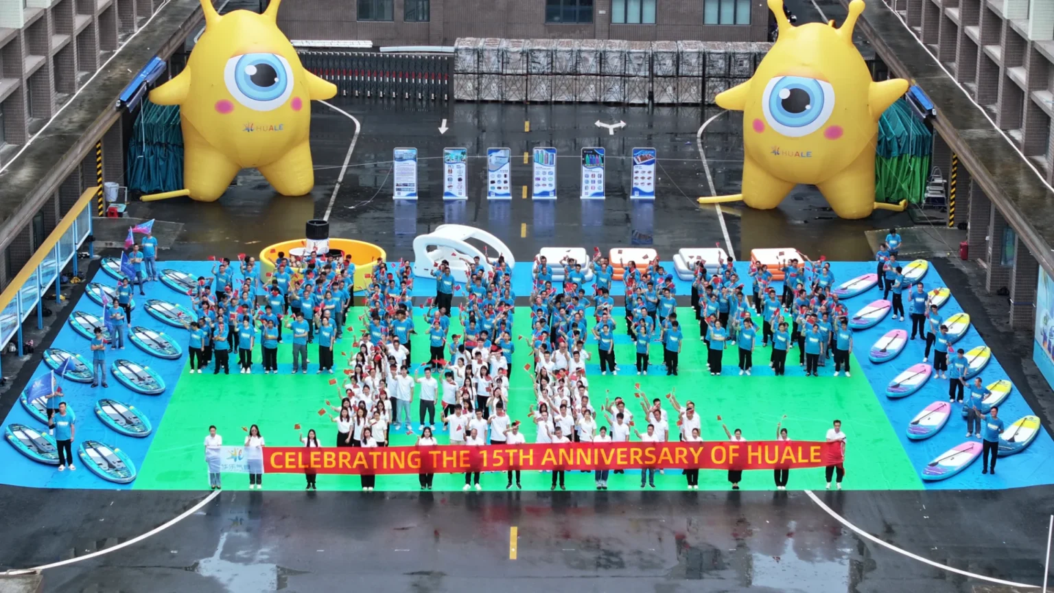 A large group of people in blue and white shirts celebrates Huale's 15th anniversary, holding flags and a banner, with inflatable yellow mascots in the background.