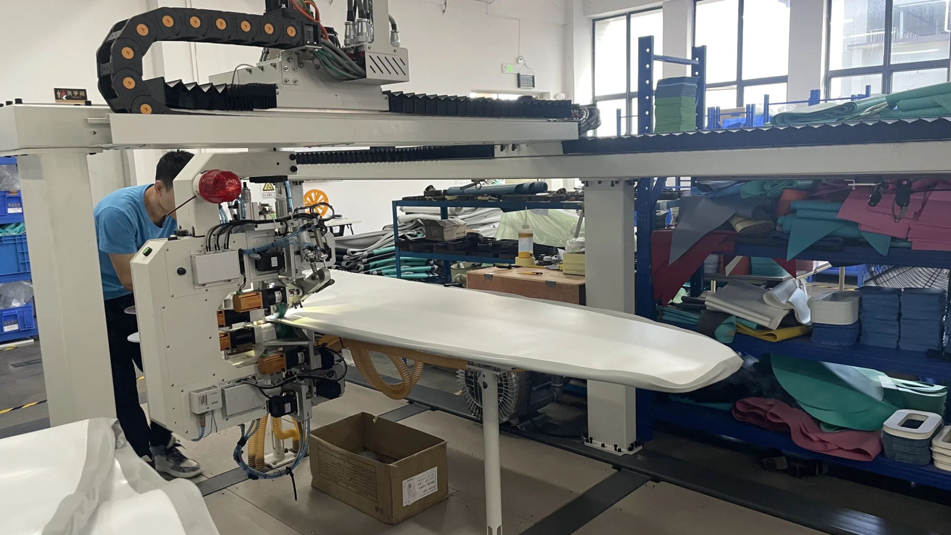 Large industrial fabric cutting machine with a white fabric being processed, operator in blue shirt adjusting machine settings, shelves with colorful materials in the background.