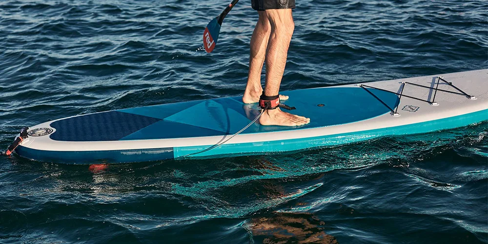 Man standing on a blue and white stand-up paddleboard on calm water, wearing a leg leash and holding a paddle.
