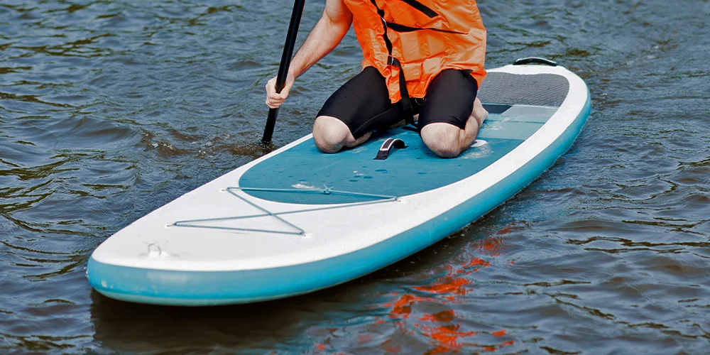 A person kneeling on a paddleboard, wearing a bright orange life vest and holding a paddle on a wavy body of water.