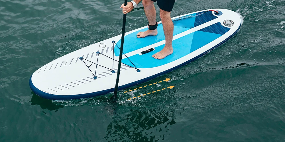 A person with shorts stand-up paddleboarding on calm water, maneuvering an inflatable board with a single paddle.