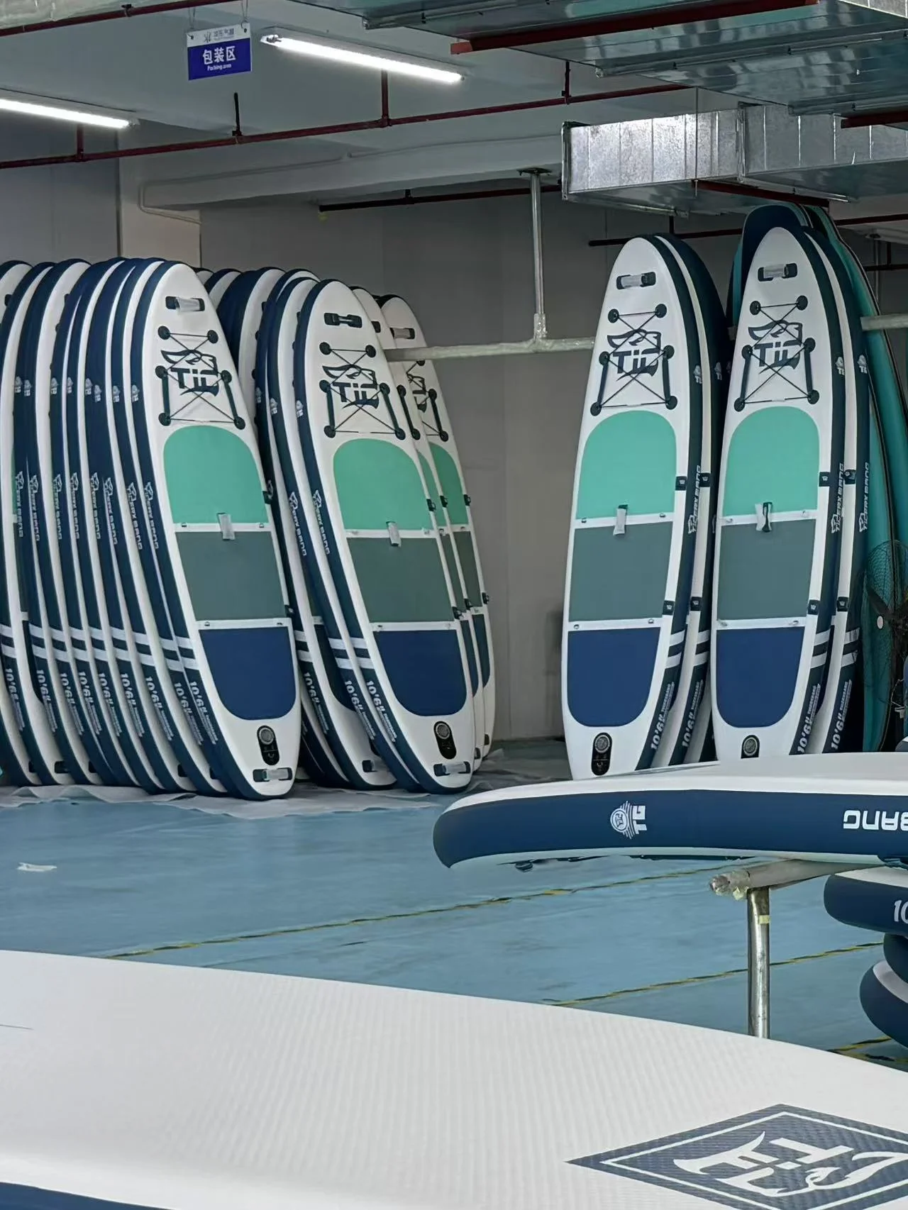 Several paddle boards stacked against a wall in a storage area, featuring teal and blue designs with visible straps and branding.