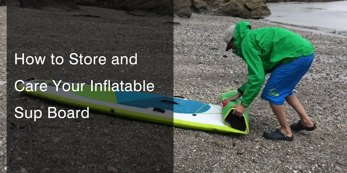 Person in green jacket and blue shorts rolling up an inflatable SUP board on a rocky beach.