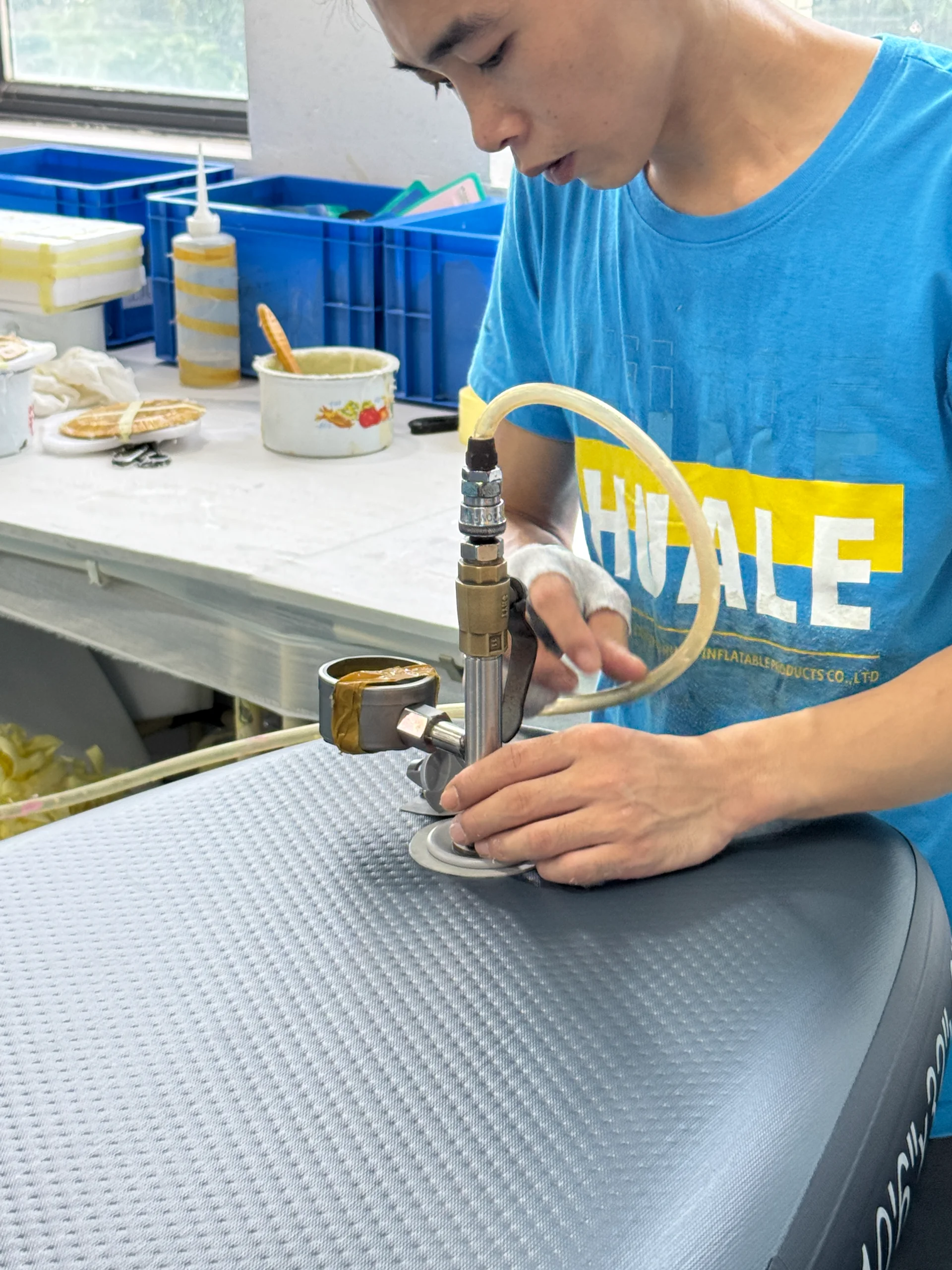 A person wearing a blue shirt operates a hand tool with a hose on textured fabric in a workshop setting, surrounded by containers and tools.