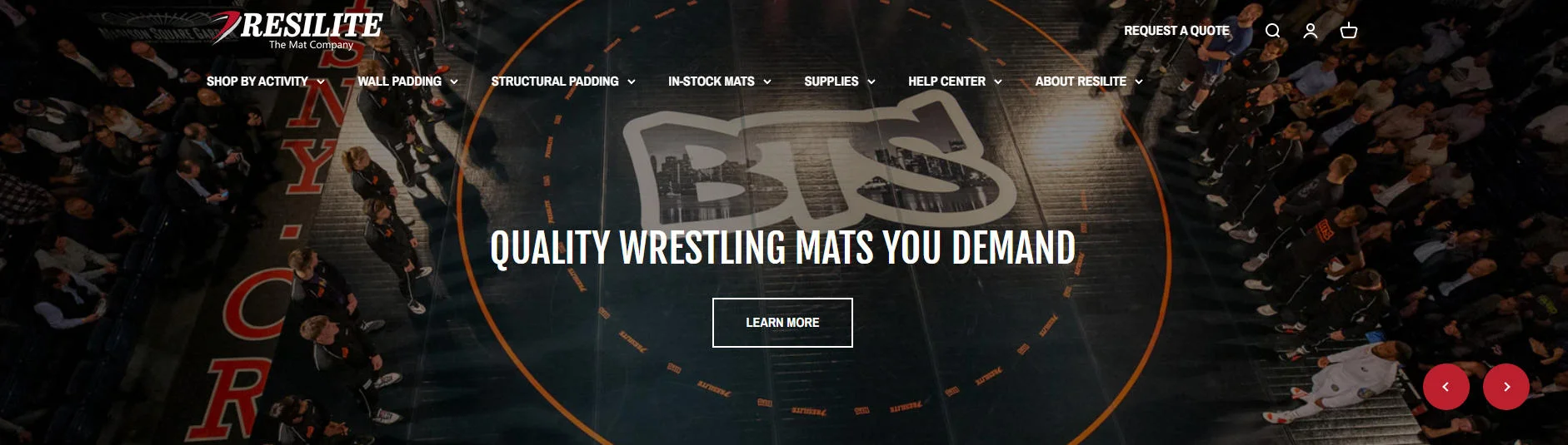 Wrestling mat event with audience in darkened arena, featuring a central mat with prominent branding and people standing around.