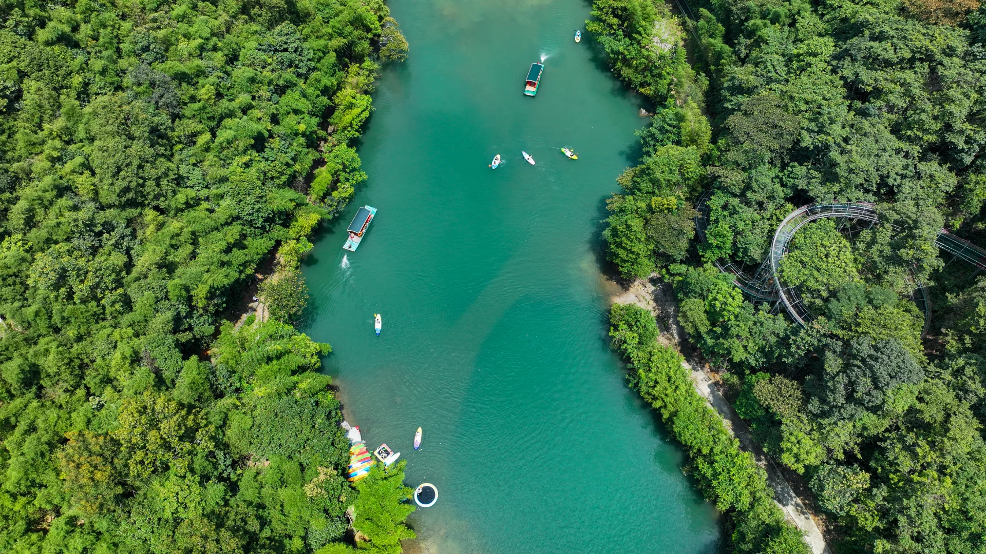 Aerial view of a blue-green river surrounded by dense forest. Small boats glide along the water, creating a serene natural scene.