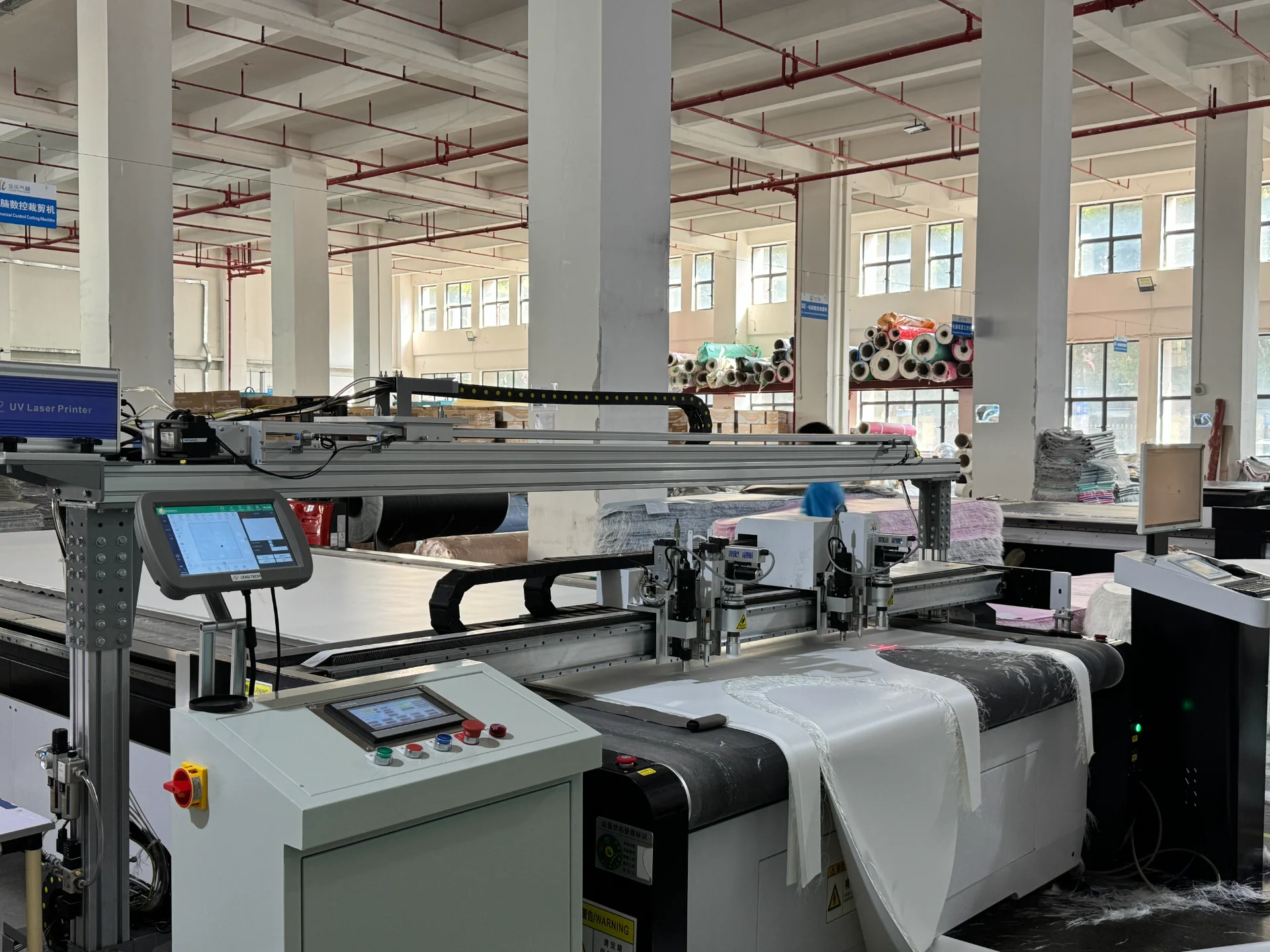 Automated laser cutting machine processing fabric in a factory setting with neatly stacked fabric rolls and large windows in the background.