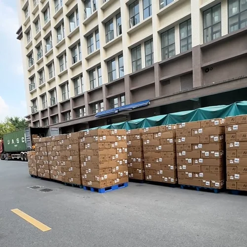 Tall stacks of sealed cardboard boxes on blue pallets outside a large warehouse building, with a truck in the background.
