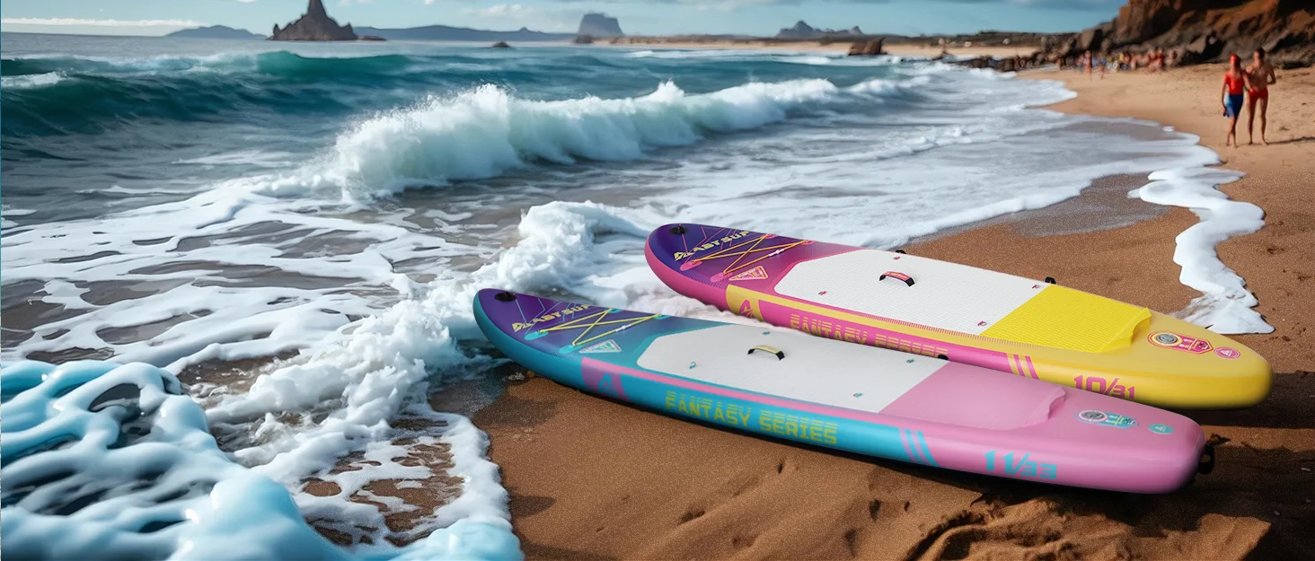 Two colorful paddleboards, one pink and one yellow, rest on a sandy beach with foamy waves nearby. Scenic coastline in the background.