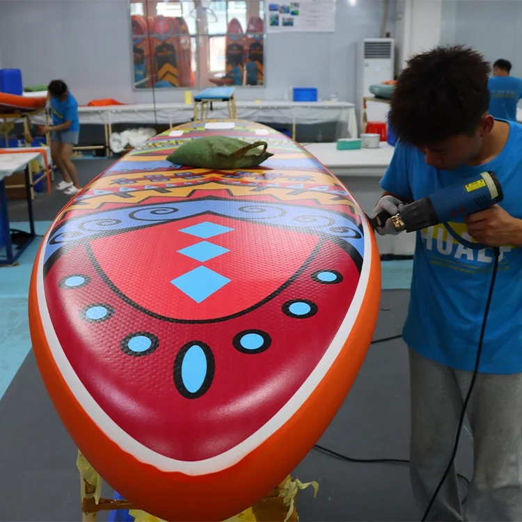 A colorful paddleboard with intricate designs being made in a workshop, with a person using a heat gun on it.