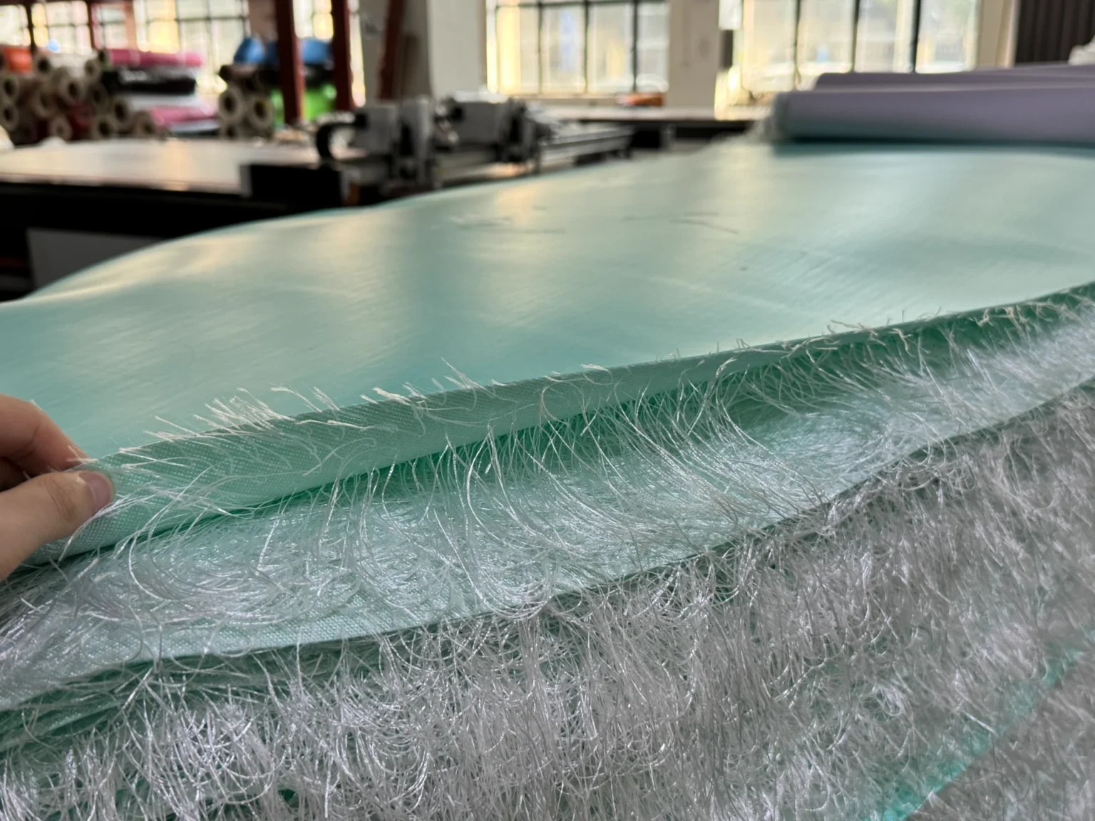 Close-up of a hand holding layers of fiberglass fabric with textured edges in a manufacturing setting, surrounded by machinery and materials.