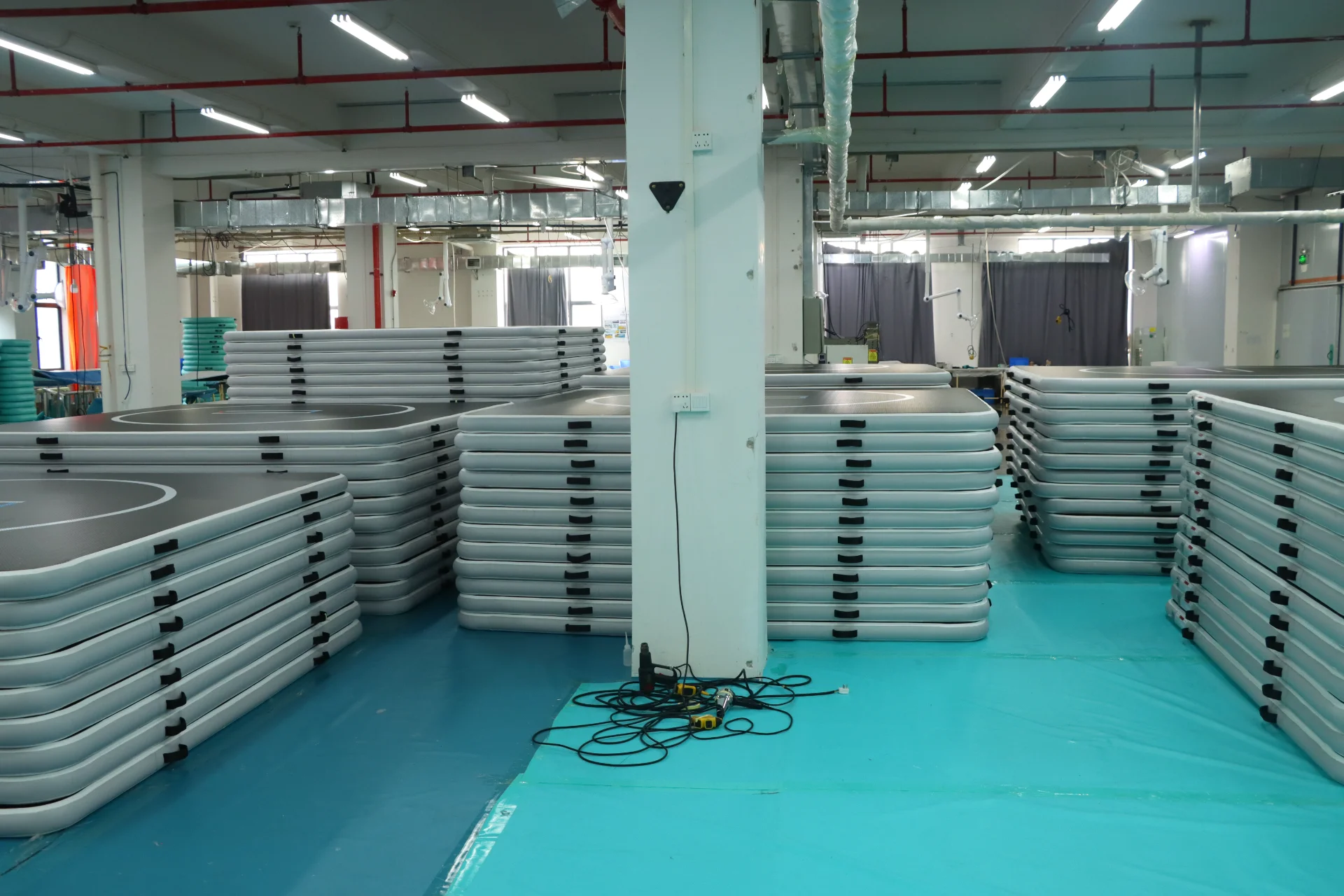 Stacks of inflatable gymnastic mats organized in a factory setting, with visible ceiling lights and industrial pipes overhead.
