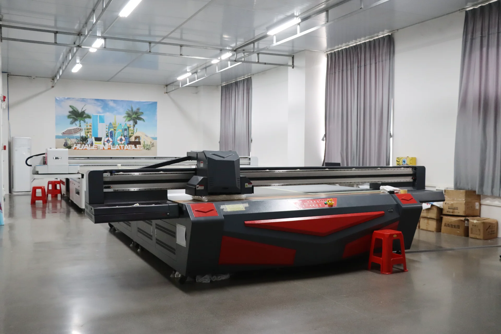 A large industrial printer with a red and black color scheme positioned in a well-lit workshop with curtains and boxes nearby.