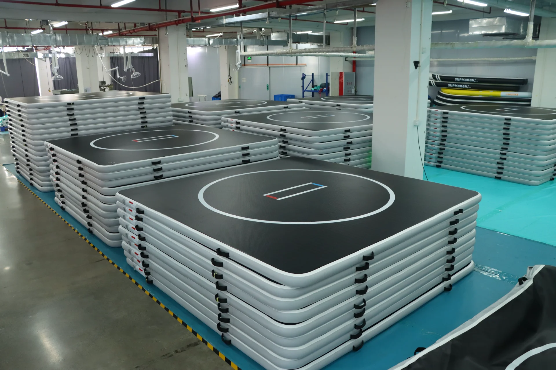 Multiple stacks of large inflatable gymnastics mats with black surfaces and white trim are neatly organized in a well-lit warehouse.