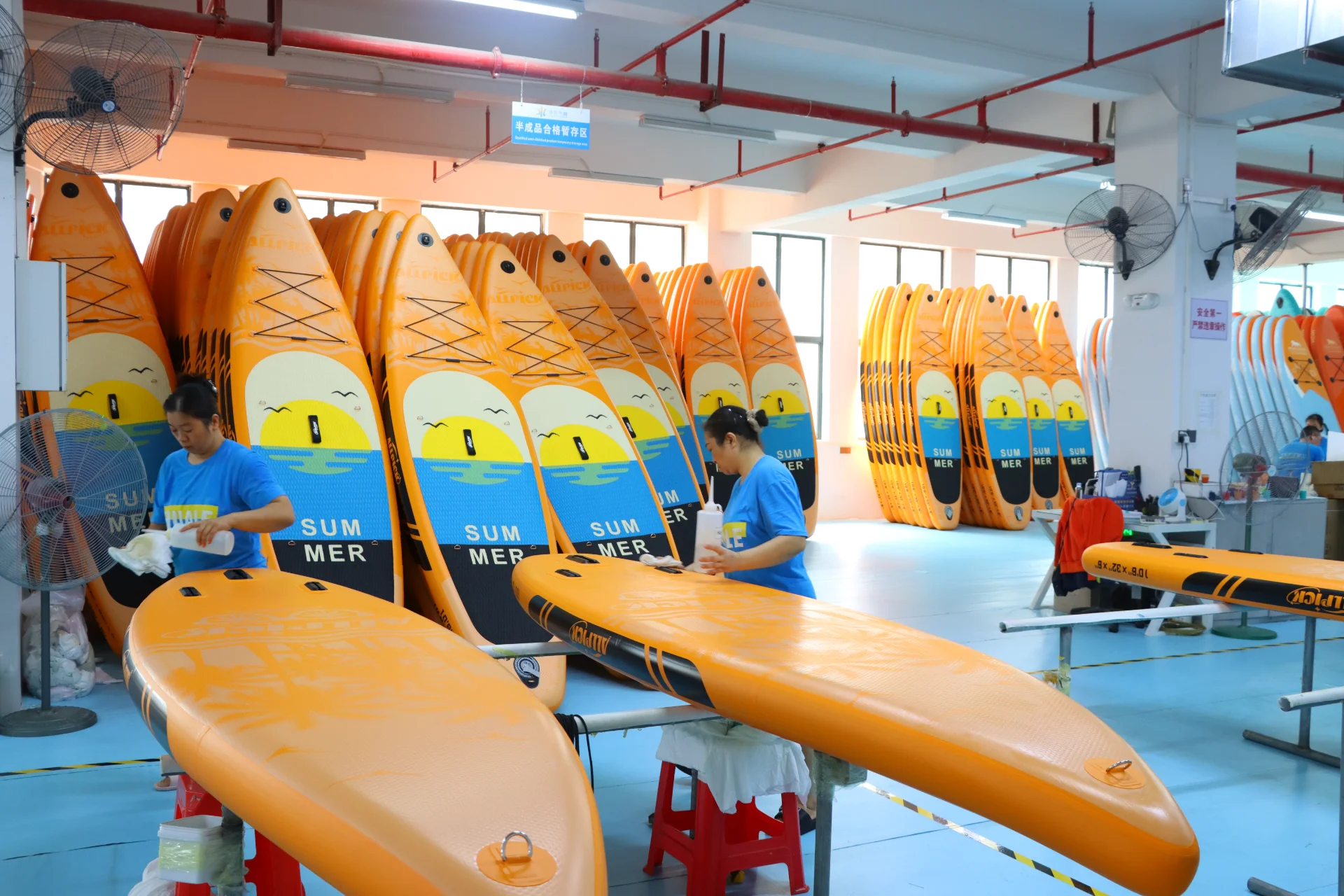 Workers assembling orange paddleboards with summer designs in a factory setting, surrounded by stacks of similar boards.