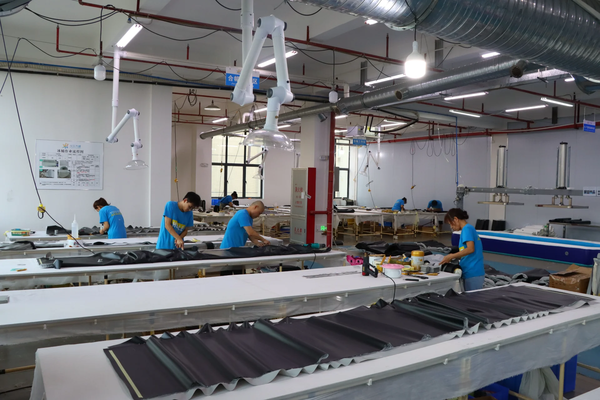 Industrial workers on a factory floor operating machinery and handling materials, surrounded by ventilation pipes and equipment.