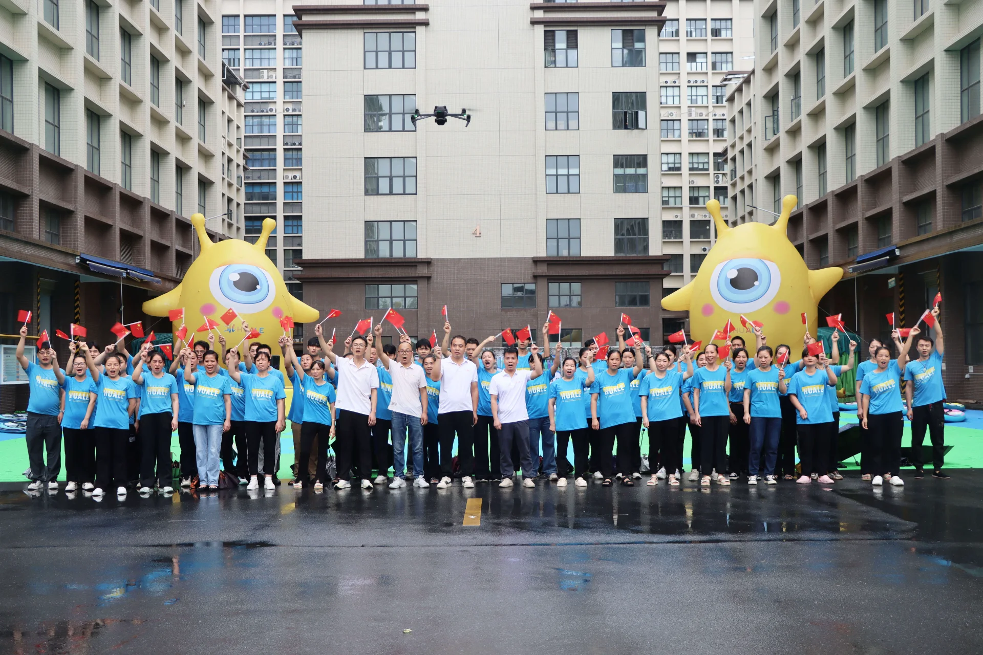 Large group of people in matching blue shirts holding small red flags, standing in front of a building with two large yellow inflatables.
