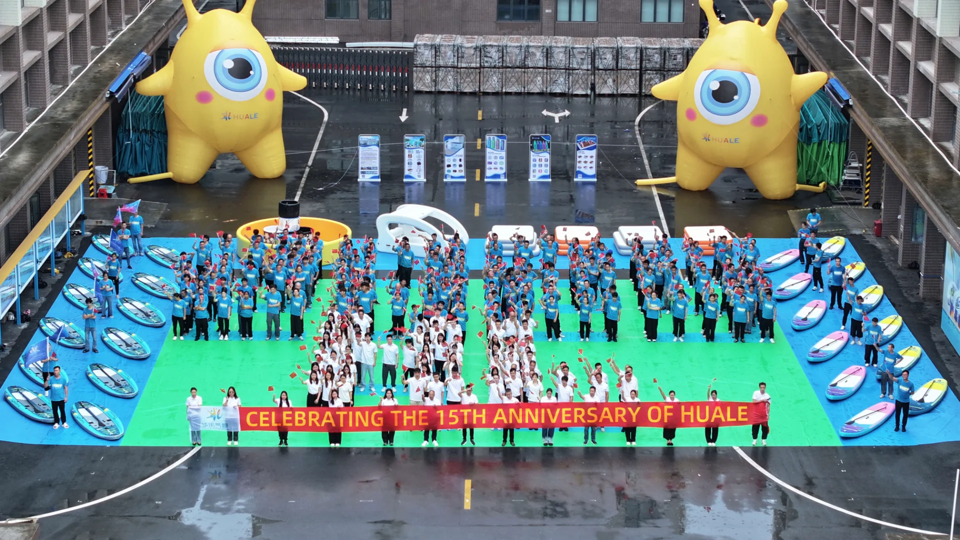 Large outdoor gathering celebrating Huale's 15th anniversary with a group of people holding a banner, inflatable structures, and decorations.