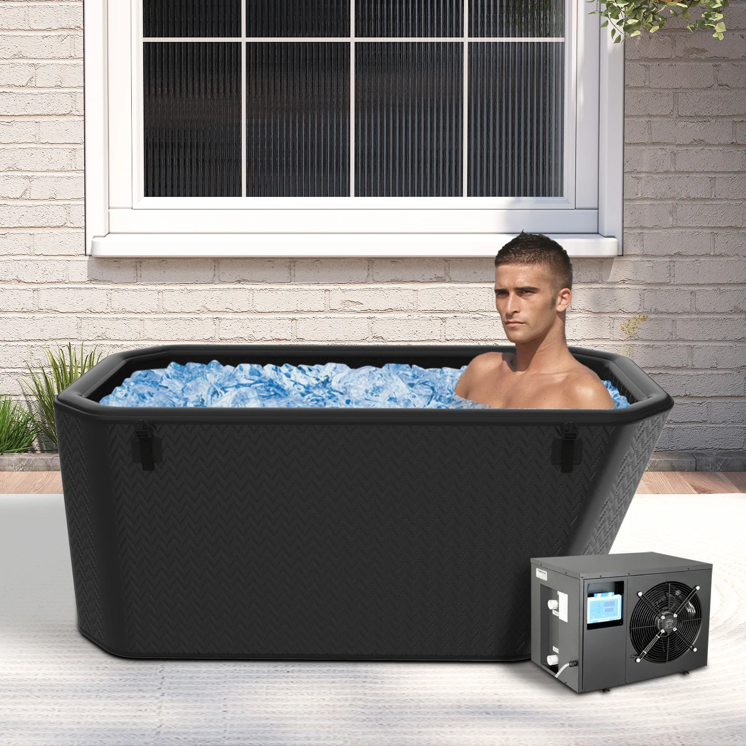 A man relaxing in a black outdoor ice bath filled with ice cubes, situated near a brick wall under a window.