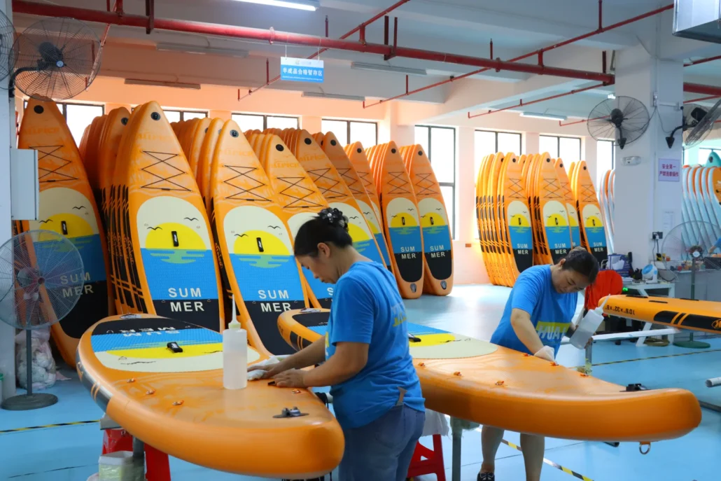 Workers assembling orange surfboards with sunset design in a factory setting.