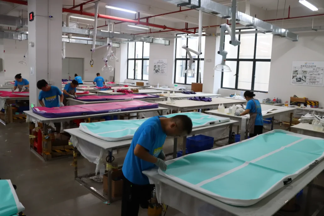 Multiple workers in a factory are crafting surfboard blanks with various colored covers. The room is well-lit with large windows.