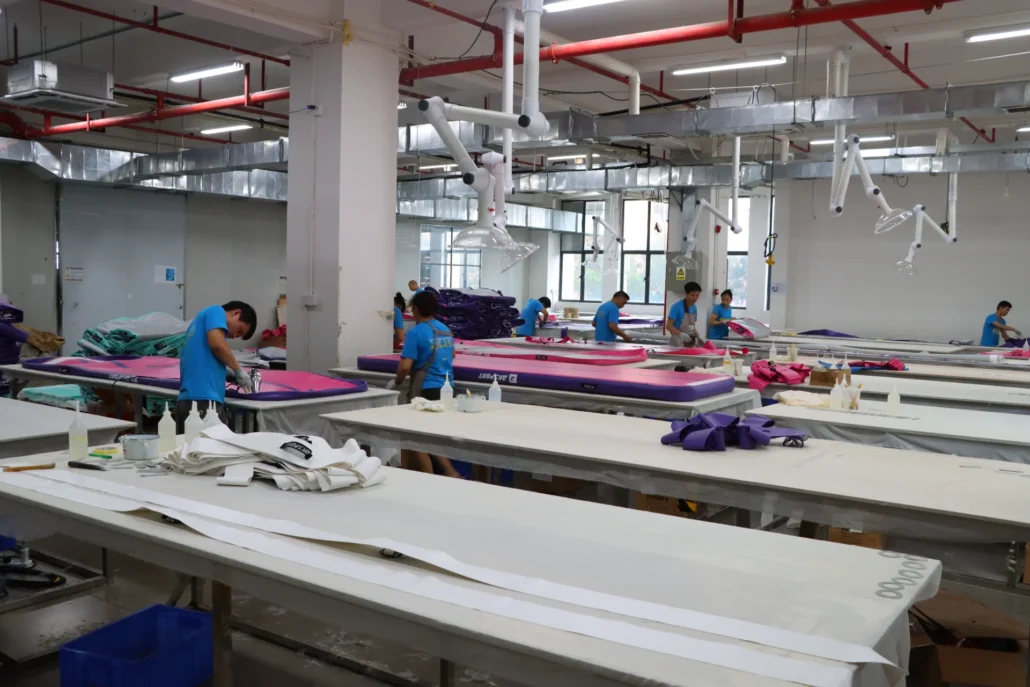 Factory workers operate on an assembly line with colorful fabric and equipment. The room is spacious and well-lit with visible ventilation.