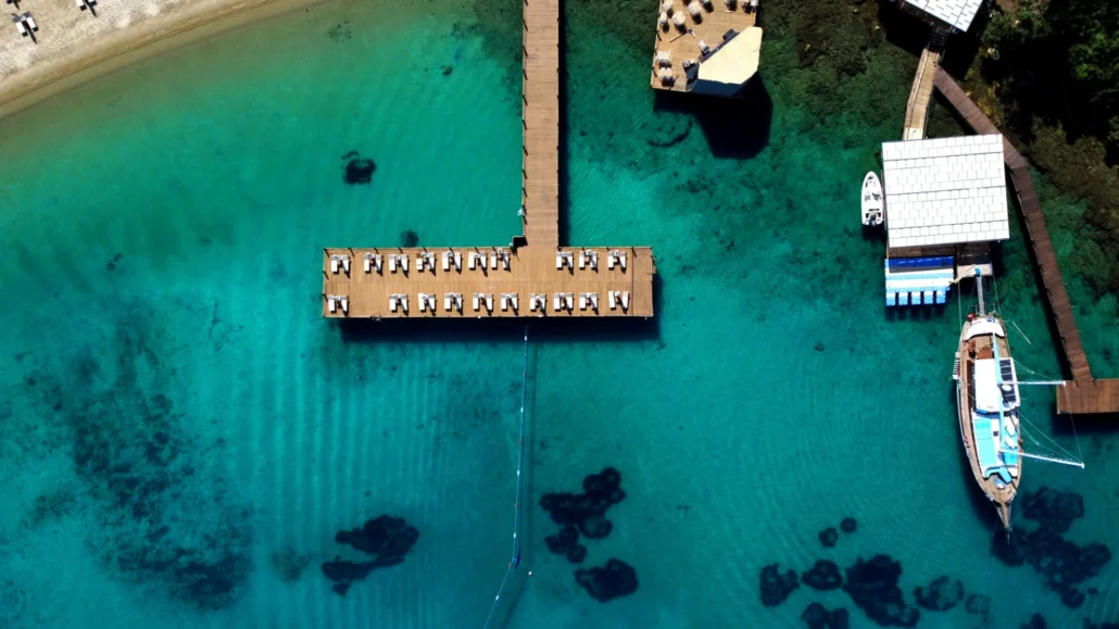 Aerial view of a wooden dock with sun loungers extending over clear turquoise water, near a beach and anchored boats.