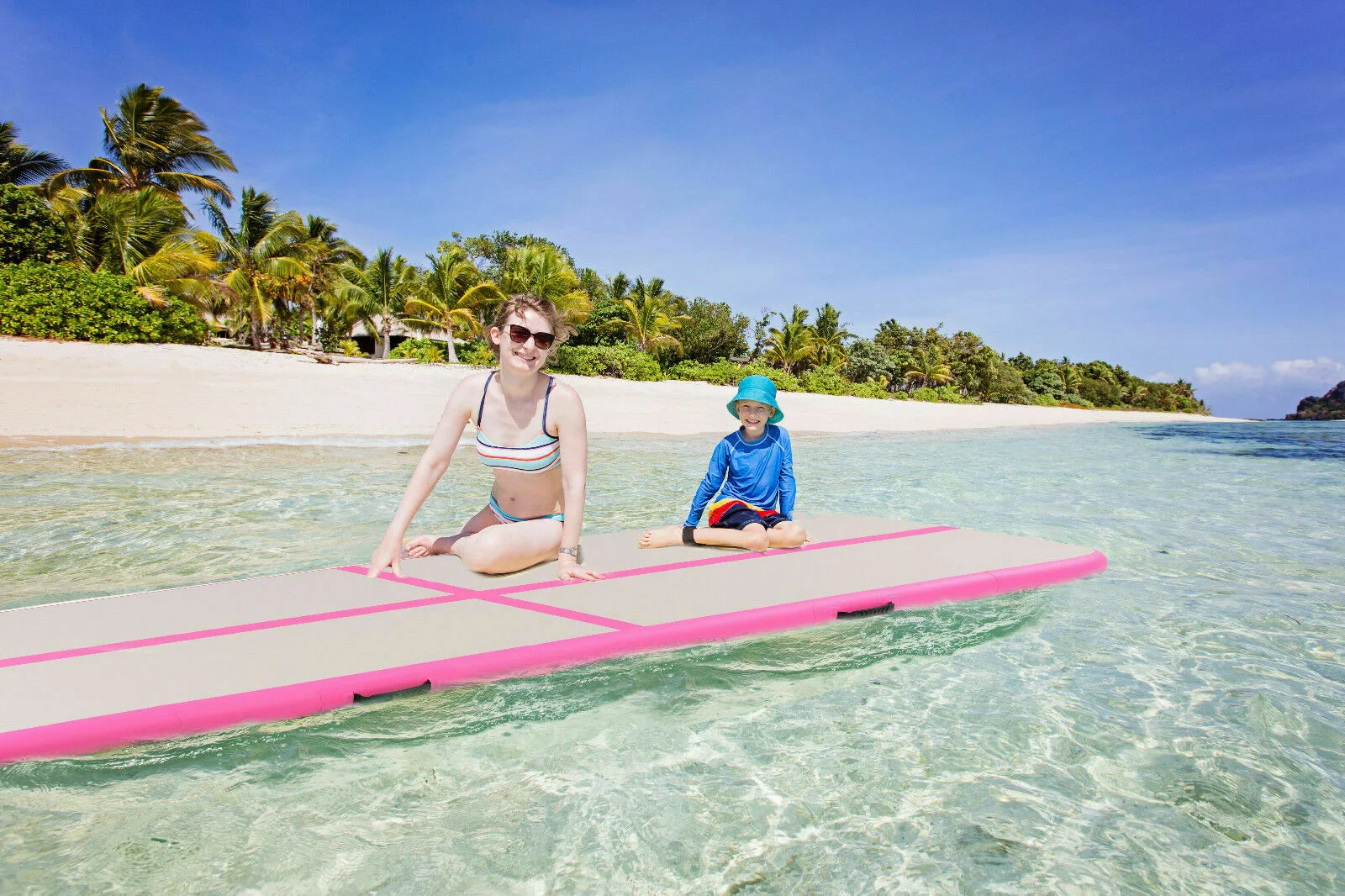 Two people sitting on a pink paddleboard on clear water near a tropical beach with palm trees under a bright blue sky.
