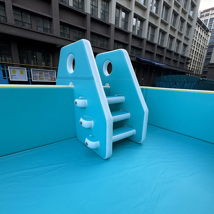 Blue playground climbing structure with steps and circular cutouts set within an enclosed area, with buildings in the background.