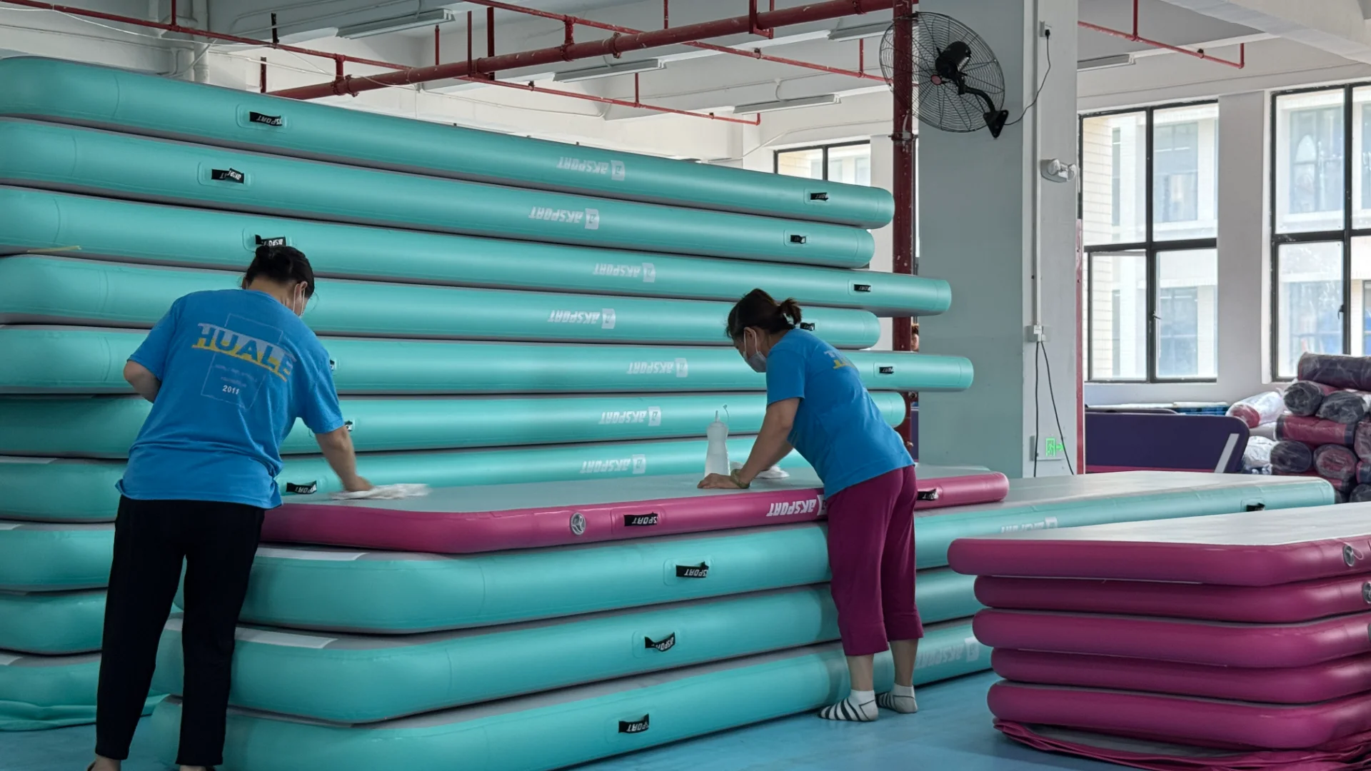 Two workers clean inflatable gymnastics mats stacked in a factory setting. Various teal and pink mats are visible, with windows and equipment in the background.