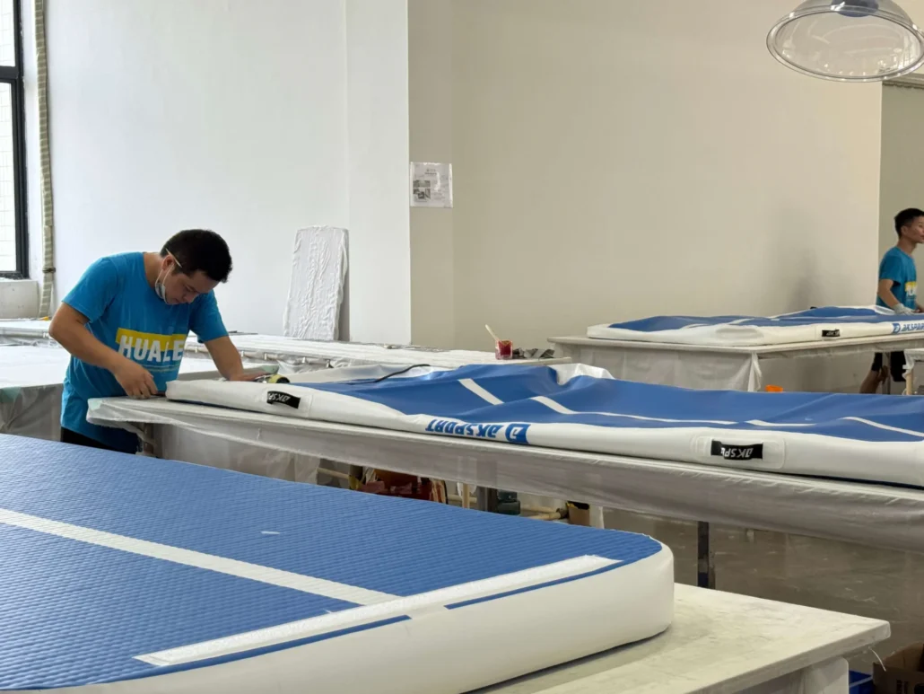 Worker assembling inflatable paddleboards on a table in a factory setting, wearing a blue Huale shirt.