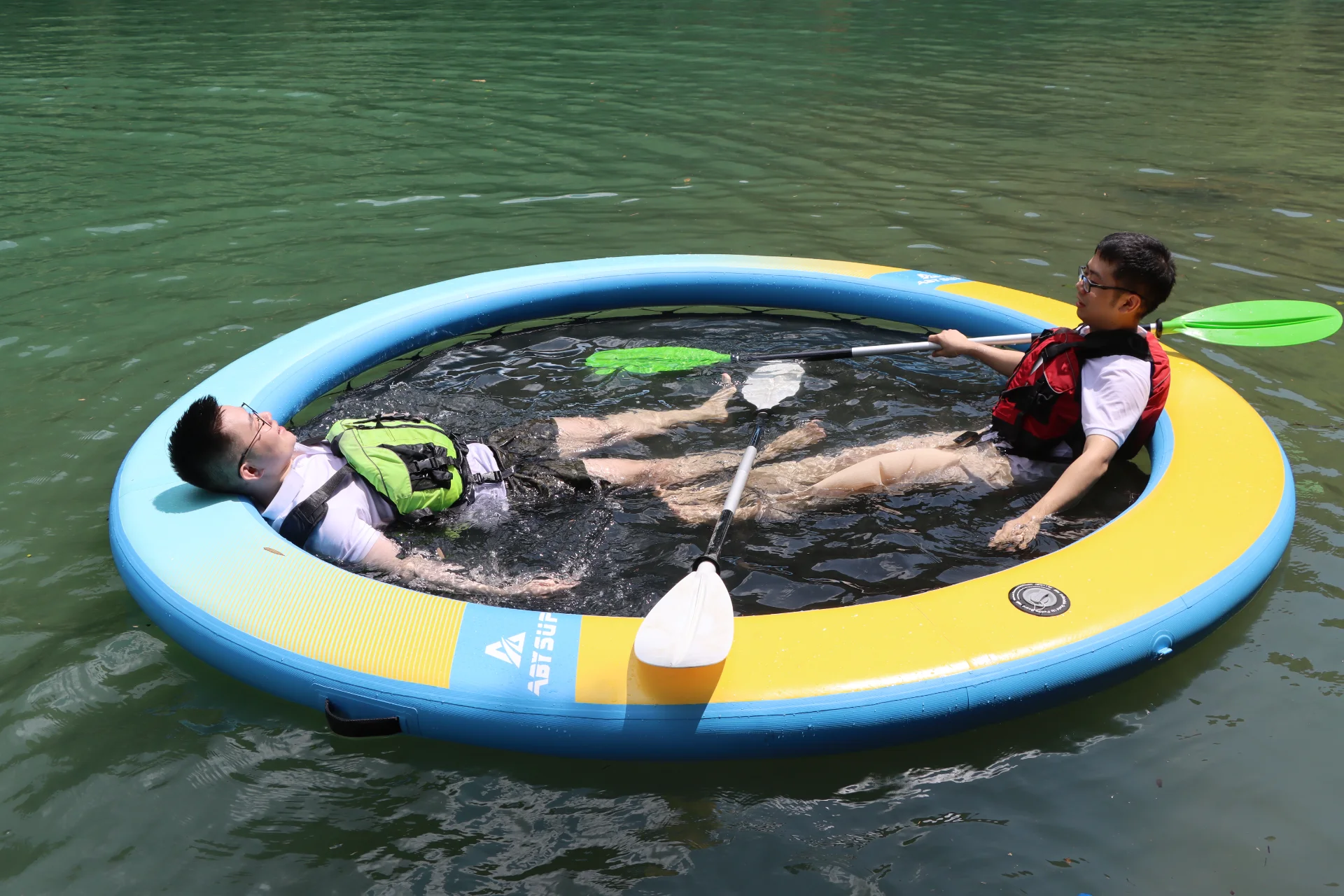 Two individuals in life vests relaxing on a large floating ring on a water surface, one holding a paddle.