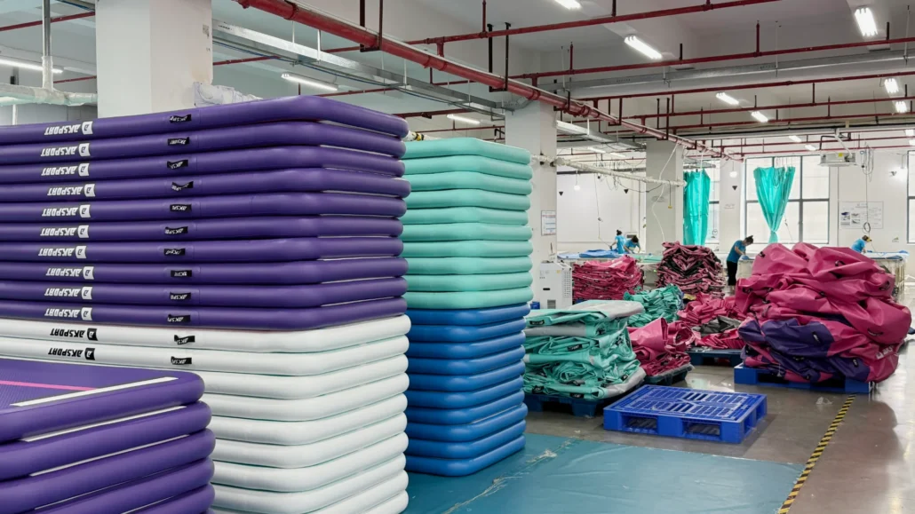 Stacks of purple, blue, and teal inflatable mats in a factory production area with workers and equipment visible in the background.
