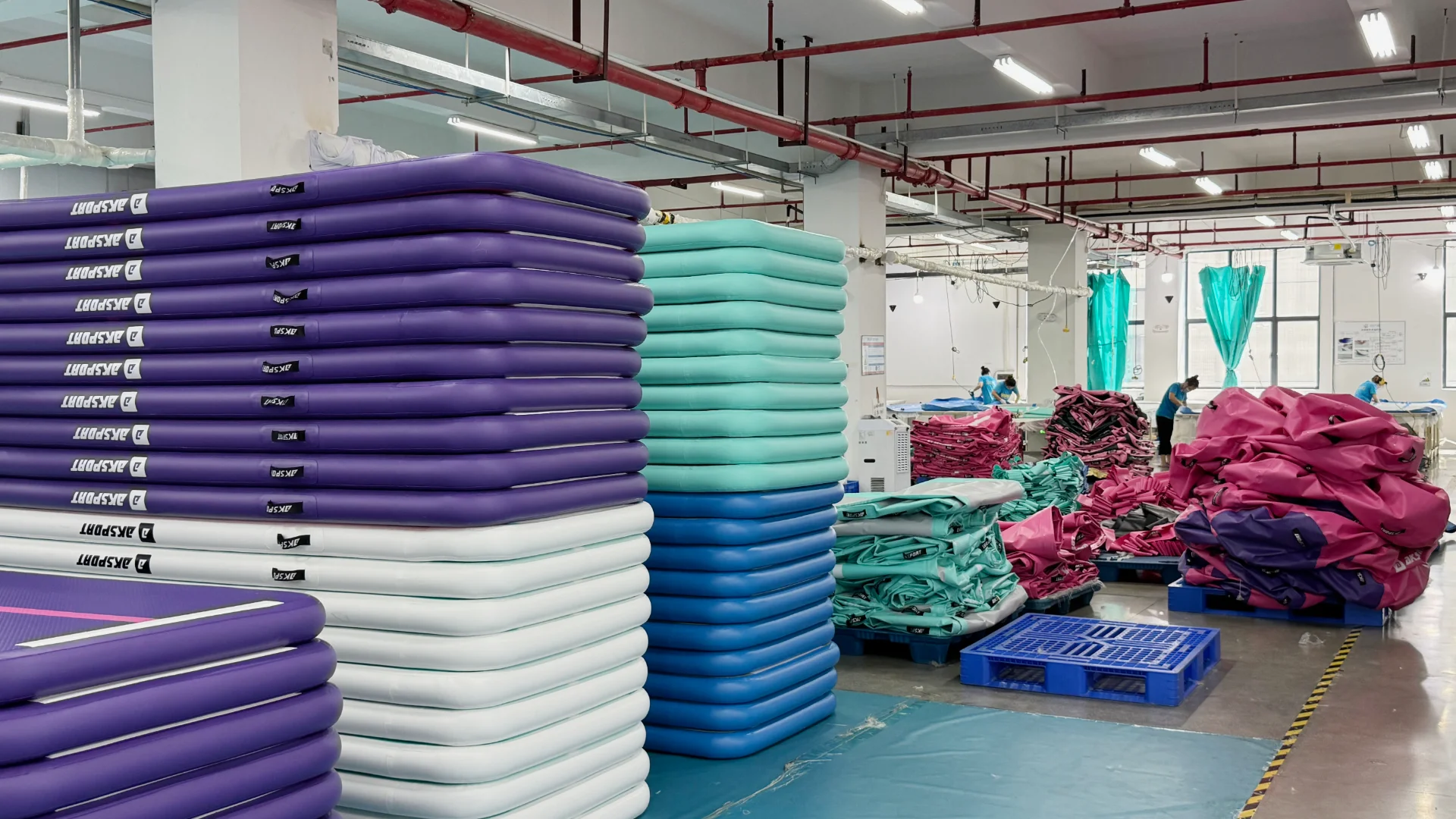 Stacks of purple, blue, and teal inflatable mats in a factory production area with workers and equipment visible in the background.