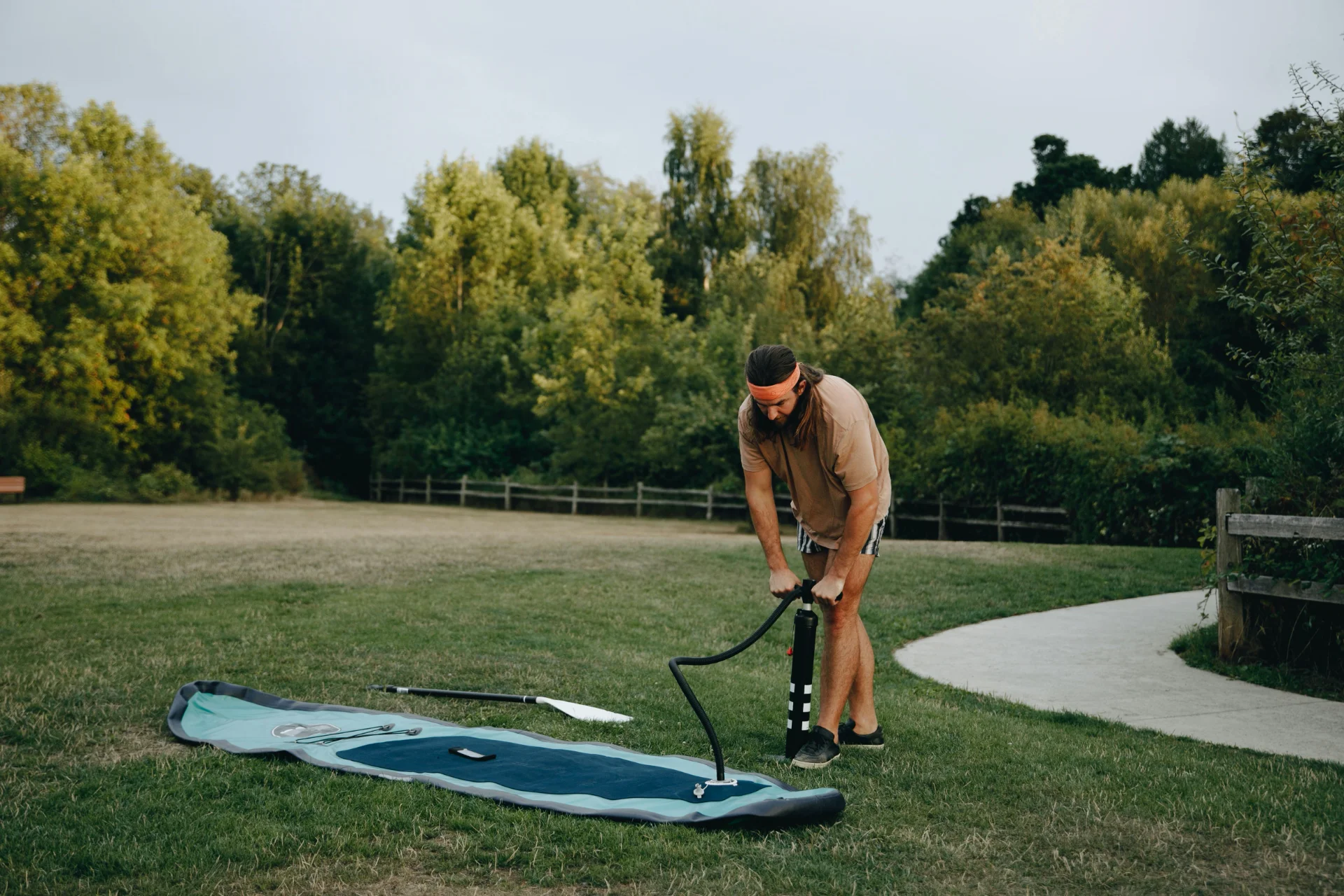 Man inflating a paddleboard with a pump in a grassy park area surrounded by trees.