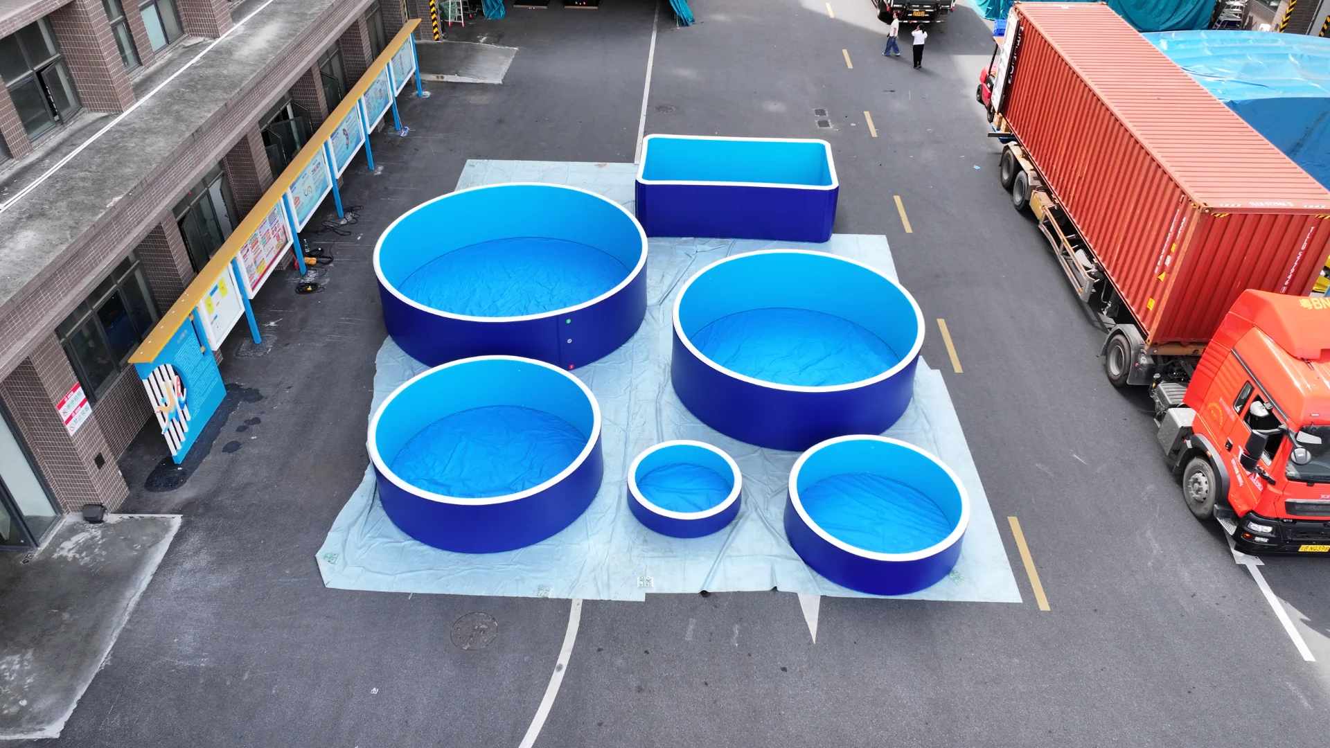 Six large, round blue pools of varying sizes arranged outdoors near an industrial building with trucks parked nearby.
