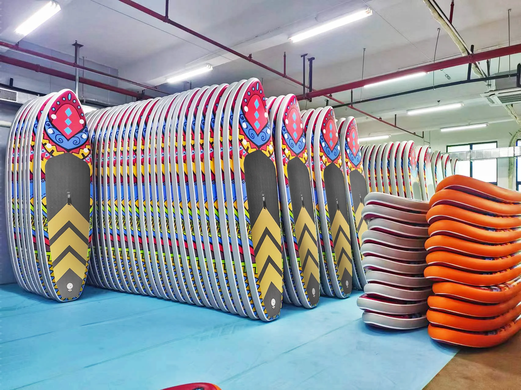Stack of colorful surfboards with vibrant designs arranged neatly in a factory setting with industrial lights overhead.