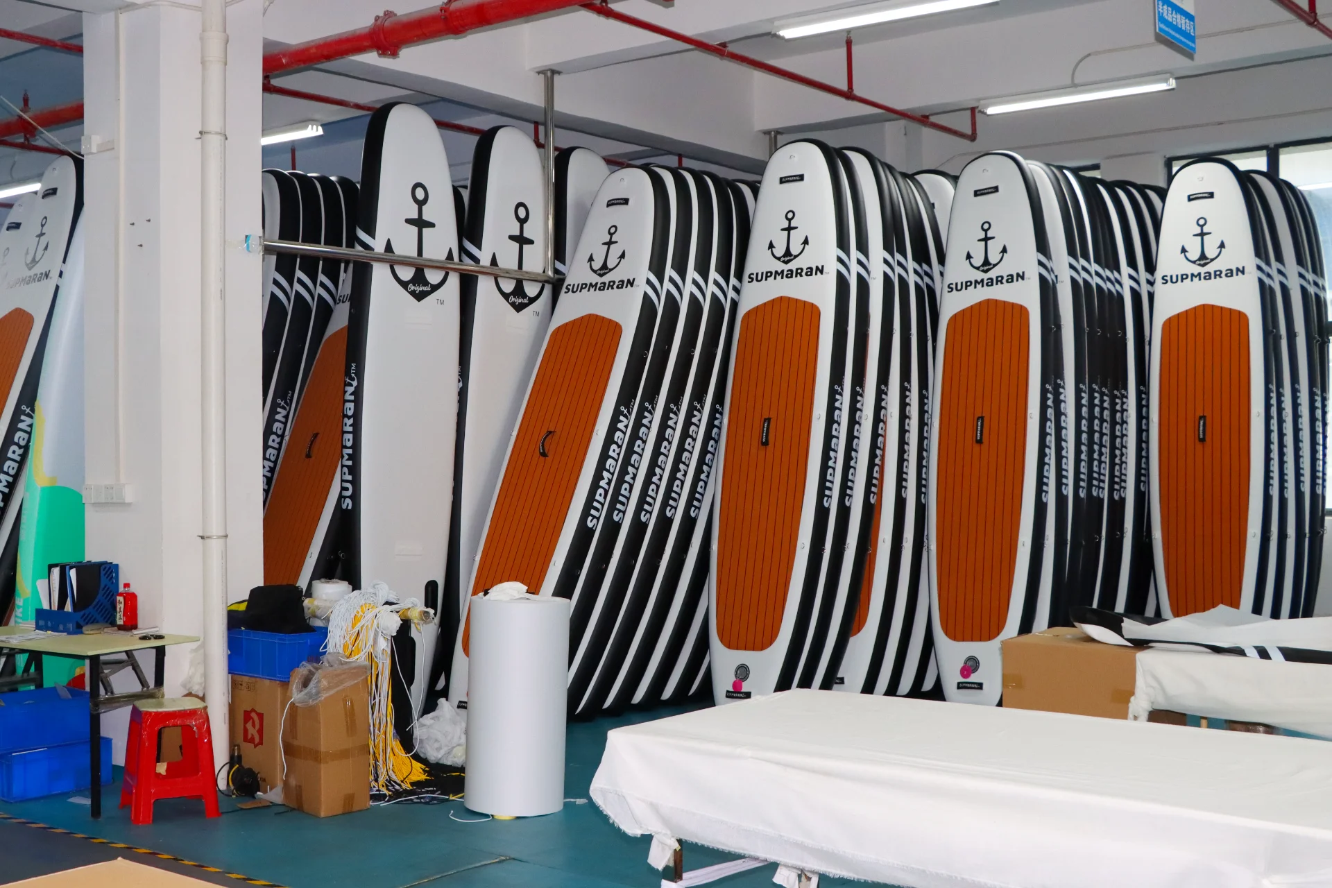 A row of paddleboards stacked vertically in a factory with visible packaging materials and tables nearby.