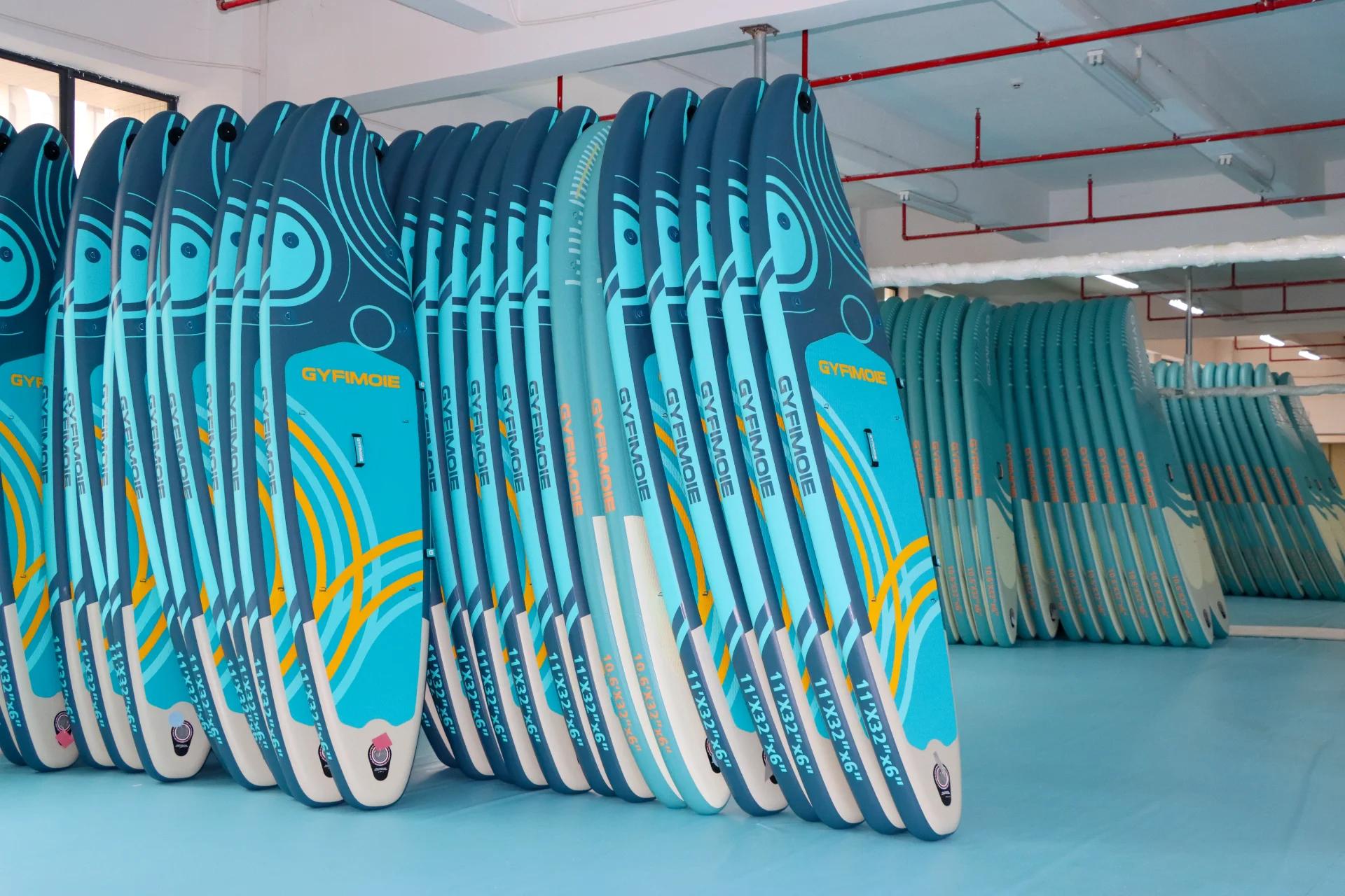 Several blue paddleboards branded GYFIMOIE are neatly stacked in a workshop, showcasing vibrant designs and sizes.