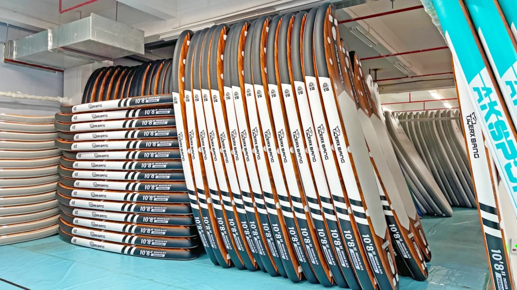 A row of paddleboards neatly stacked in a storage room with visible labels and vibrant designs.