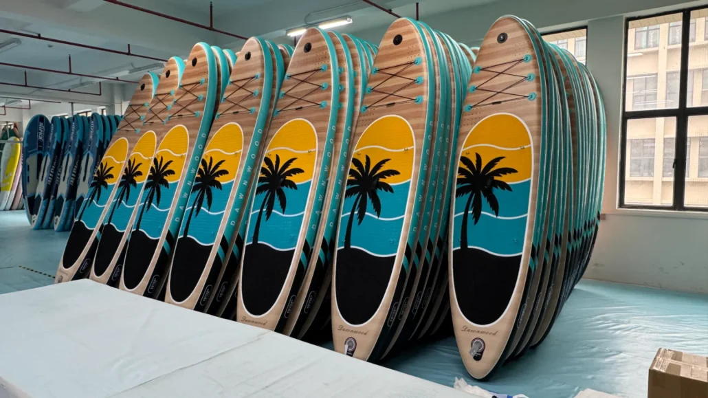 Several surfboards with palm tree designs stacked vertically in a factory building, with windows letting in natural light.