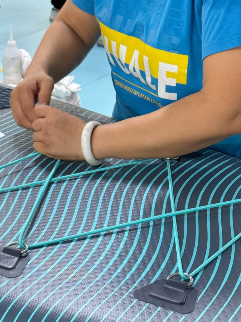 A person in a blue shirt assembling an inflatable paddleboard with turquoise lines and bungee cords.