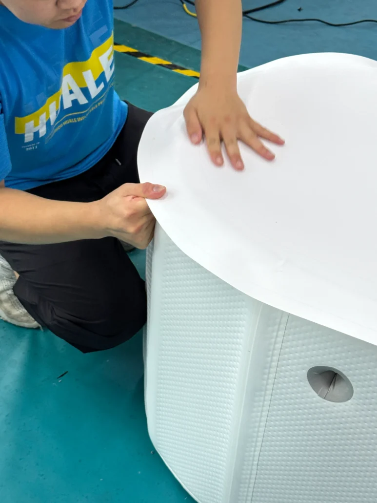 A worker wearing a blue shirt assembles a white plastic component with textured sides on a blue floor.