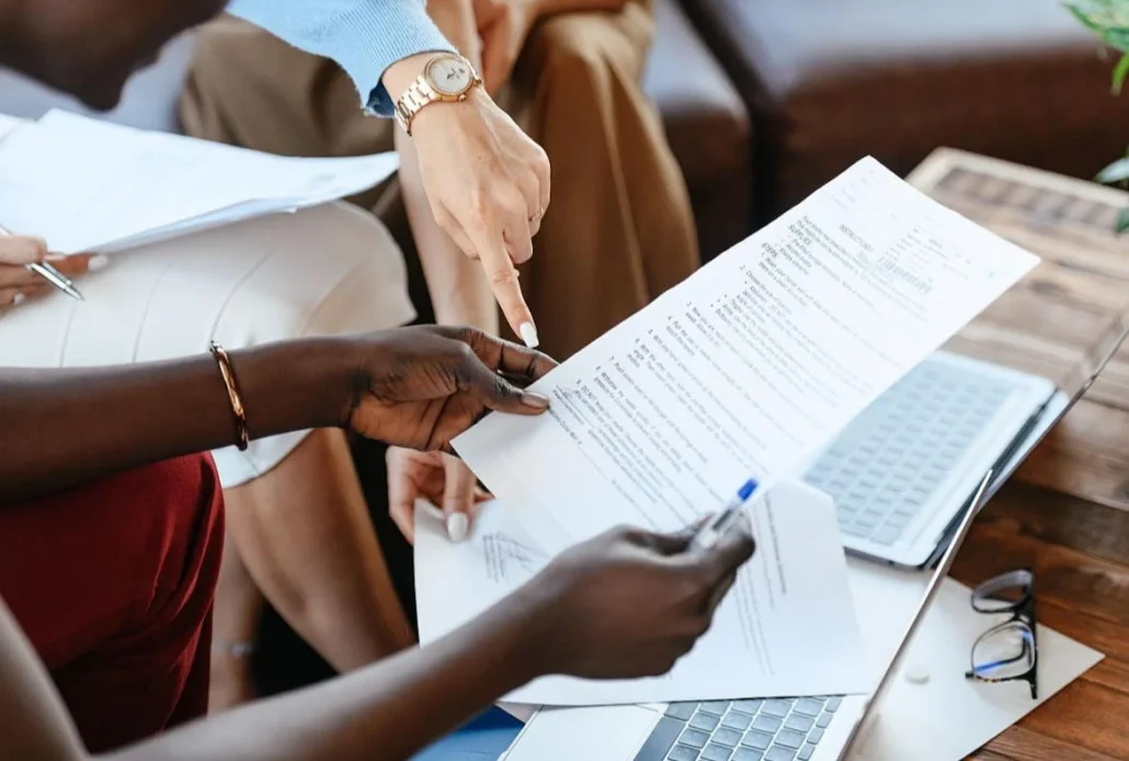 Group of people in a business meeting examining a printed document, with a laptop and glasses on the table.