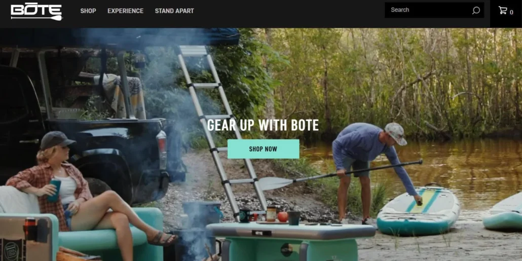 Outdoor camping setup by a riverside with BOTE gear, featuring a woman relaxing and a man preparing a paddleboard.
