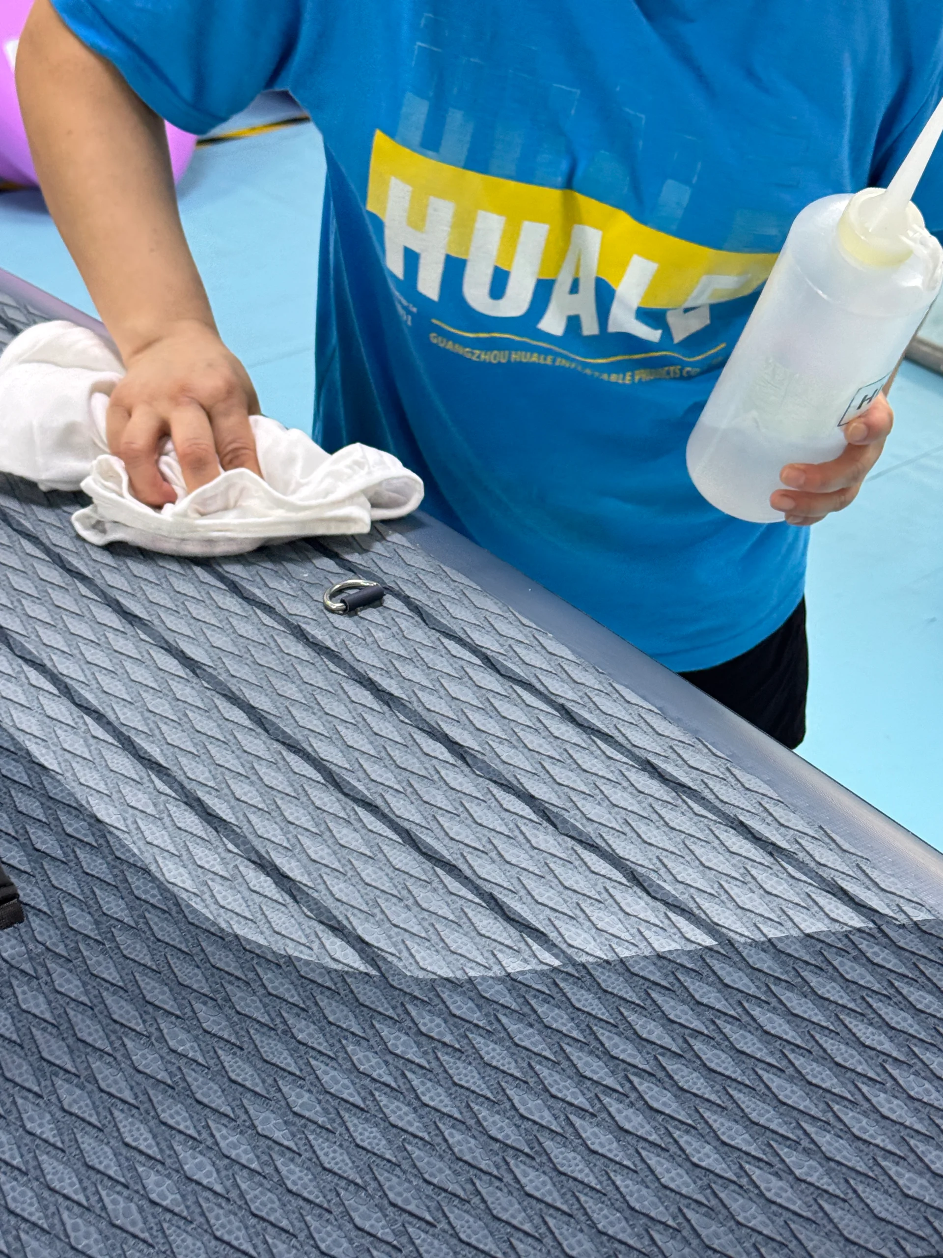 Person in a blue shirt cleaning a gray inflatable paddleboard with a cloth and spray bottle, showing a textured surface.